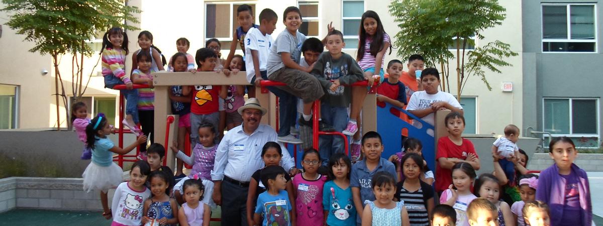 A large group of children and Tony Salazar gather around a playground at MacArthur Park Apartments